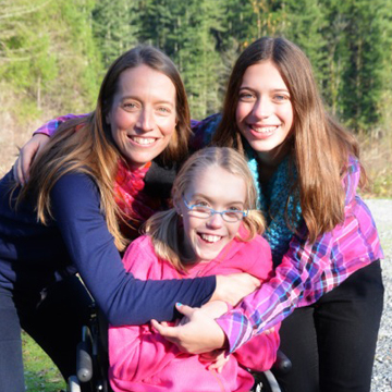 Girl in wheelchair smiling and being hugged by her mom and sister